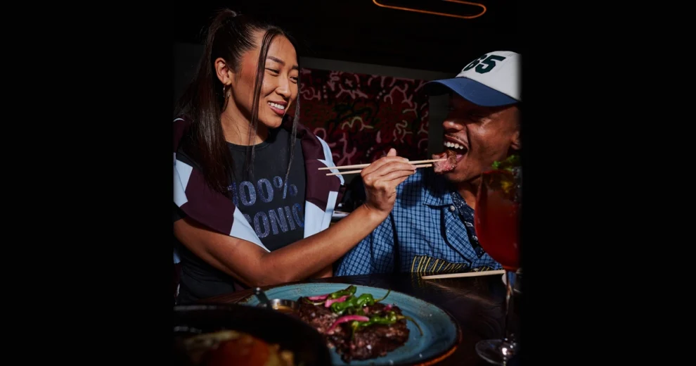 A diner at KYU Miami smiles while using chopsticks to feed a bite of grilled meat to a companion seated beside them. A plated grilled entrée and a cocktail are visible on the table, highlighting the restaurant's lively, social dining atmosphere during Miami Spice Months.