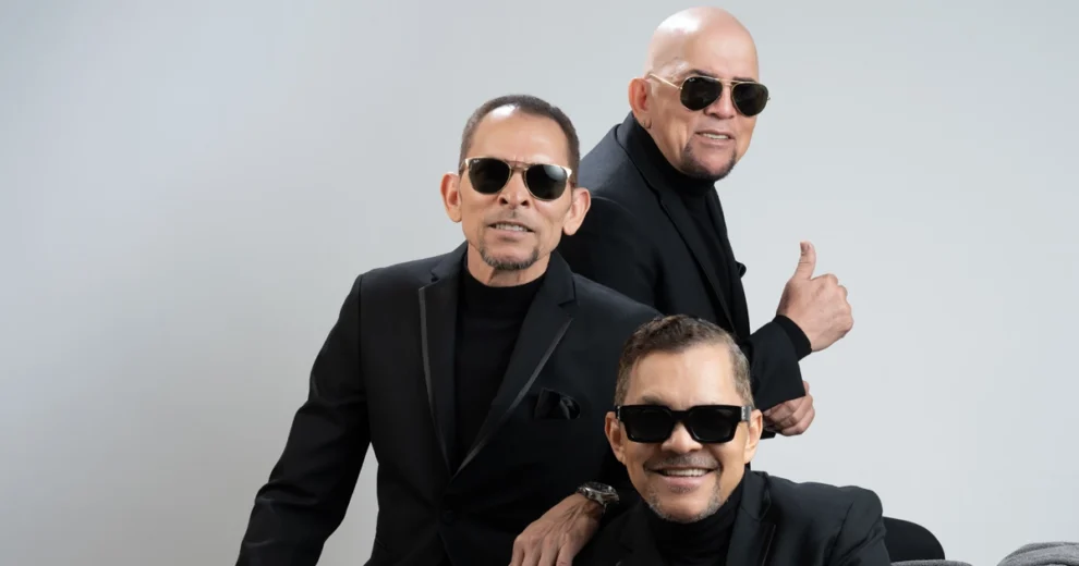 Tres miembros de Los Hermanos Rosario posan juntos en un estudio, frente a un fondo gris claro. Los tres visten trajes negros con suéteres de cuello alto negros y llevan gafas de sol oscuras.