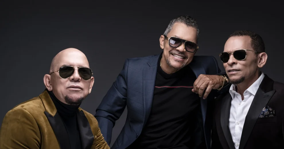 Three members of Los Hermanos Rosario pose together in a studio against a dark background.