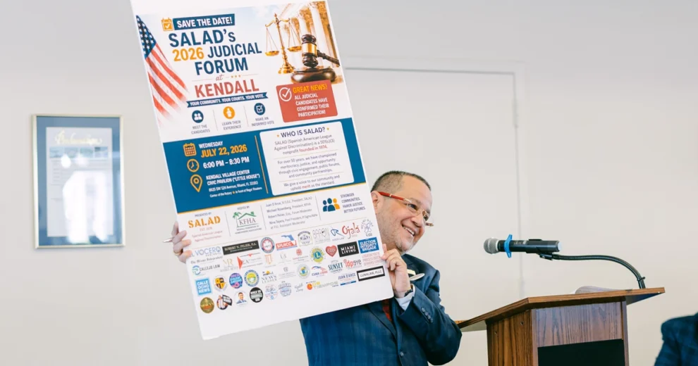 Un hombre con traje está de pie ante un podio y sostiene un gran cartel promocional del Foro Judicial de SALAD de 2026 en Kendall.