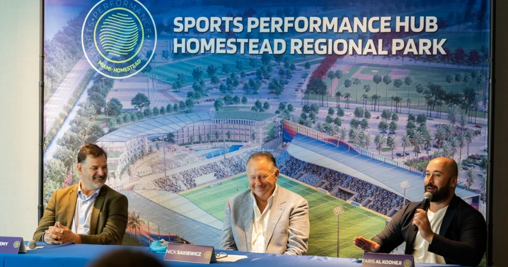 Three speakers sit at a panel table during a press conference for the Sports Performance Hub at Homestead Regional Park. A large rendering of the planned sports complex fills the backdrop as Faris Al Kooheji speaks into a microphone, with Nick Sakiewicz and Sean Remy seated beside him listening and smiling.