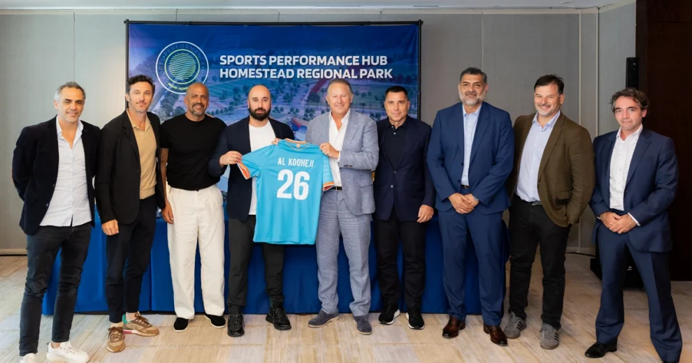 Un grupo de nueve hombres posa frente a una pancarta del Sports Performance Hub en el Homestead Regional Park durante un evento de prensa. Dos de los participantes sostienen una camiseta de fútbol de color azul claro con el nombre "Al Kooheji" y el número 26, mientras los demás se sitúan a su lado vistiendo ropa formal e informal.