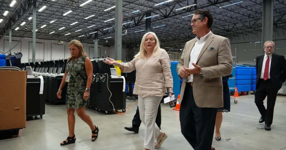 Un grupo de funcionarios electorales y visitantes recorre un almacén lleno de filas de equipos de votación durante una visita a unas instalaciones de operaciones electorales.