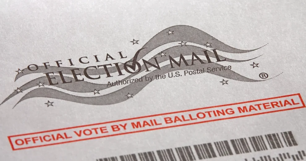 Close-up of an official Vote-by-Mail ballot envelope displaying the "Official Election Mail" logo, U.S. Postal Service authorization, and "Official Vote by Mail Balloting Material" label.