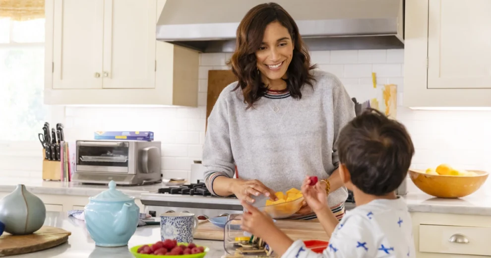 Un adulto sonriente prepara fruta fresca en una cocina luminosa, mientras un niño pequeño intenta alcanzar una pieza de fruta sobre la encimera.