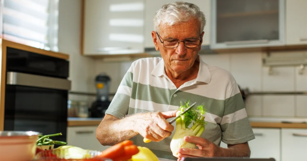 An older adult wearing glasses prepares fresh vegetables in a home kitchen, carefully trimming a fennel bulb on a cutting board surrounded by carrots and other produce. The image emphasizes healthy meal preparation and independent living through nutritious cooking.