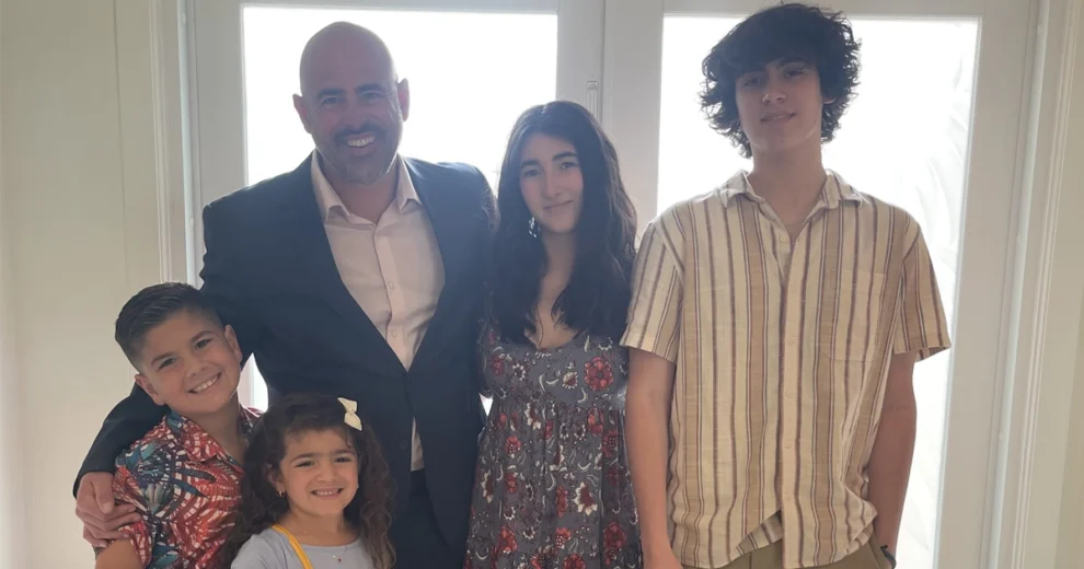 Alex Annunziato está de pie, sonriendo, junto a cuatro niños frente a unas puertas de cristal muy luminosas.