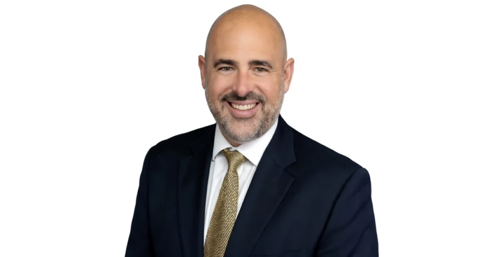Professional headshot of Alex Annunziato smiling at the camera. He is wearing a navy blue suit, a white dress shirt, and a gold patterned tie against a clean white background.