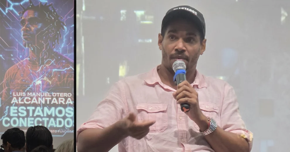 Luis Manuel Otero Alcántara habla por un micrófono dirigiéndose al público durante un coloquio sobre el documental "Luis Manuel Otero Alcántara: Estamos Conectados". Viste una camisa rosa claro y una gorra negra, y gesticula mientras se proyecta una imagen promocional de la película en una pantalla a su lado.