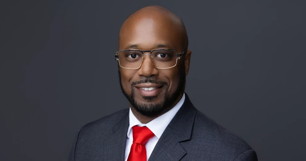Professional headshot of Arthur McNeil wearing a gray suit, white shirt, and red tie. He is smiling and wearing glasses against a dark gray studio background.