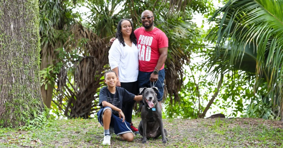 Arthur McNeil posa al aire libre con su familia en un parque de exuberante vegetación. De pie junto a su pareja, viste una camiseta roja con la inscripción "Proud Dad" (Papá orgulloso), mientras su hijo pequeño está arrodillado junto al perro gris de la familia, en primer plano.