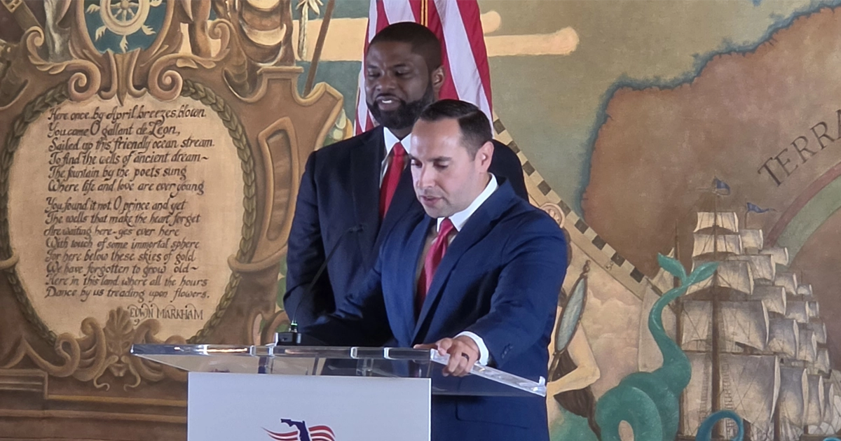 Bryan Avila speaks at a podium while Byron Donalds stands behind him. Both men wear dark suits with red ties. An American flag and a nautical-themed mural are visible in the background.