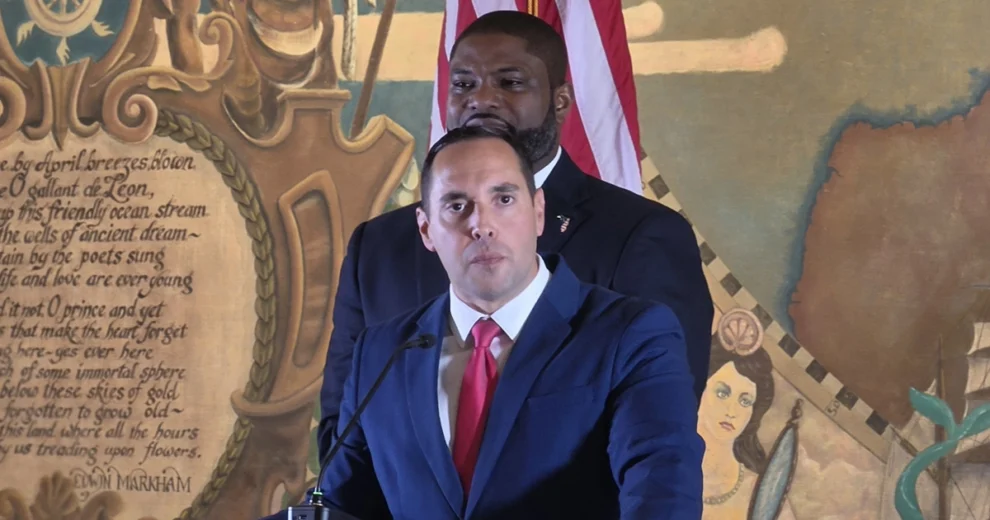 Bryan Avila está de pie ante un podio, vestido con traje azul y corbata roja, y habla por un micrófono en un evento. Byron Donalds se encuentra detrás de él; al fondo se ven una bandera de Estados Unidos y un mural colorido.