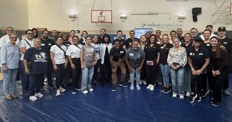 Un numeroso grupo de voluntarios, miembros del personal y participantes de la comunidad posa para una foto de grupo en el interior del gimnasio Peebles, durante un evento de regreso a clases organizado por The Caring Place.