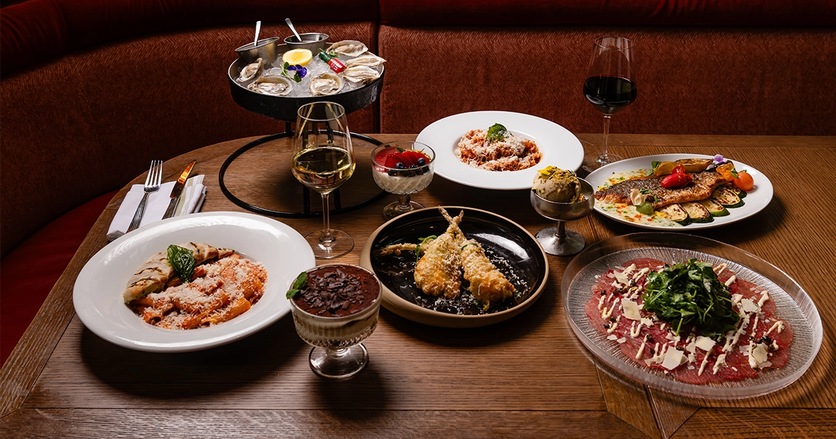 A restaurant table featuring an assortment of Italian dishes, including pasta, seafood, carpaccio, fried seafood, vegetables, desserts, oysters, and glasses of wine. The dishes are arranged on a wooden table in a warmly lit dining area.