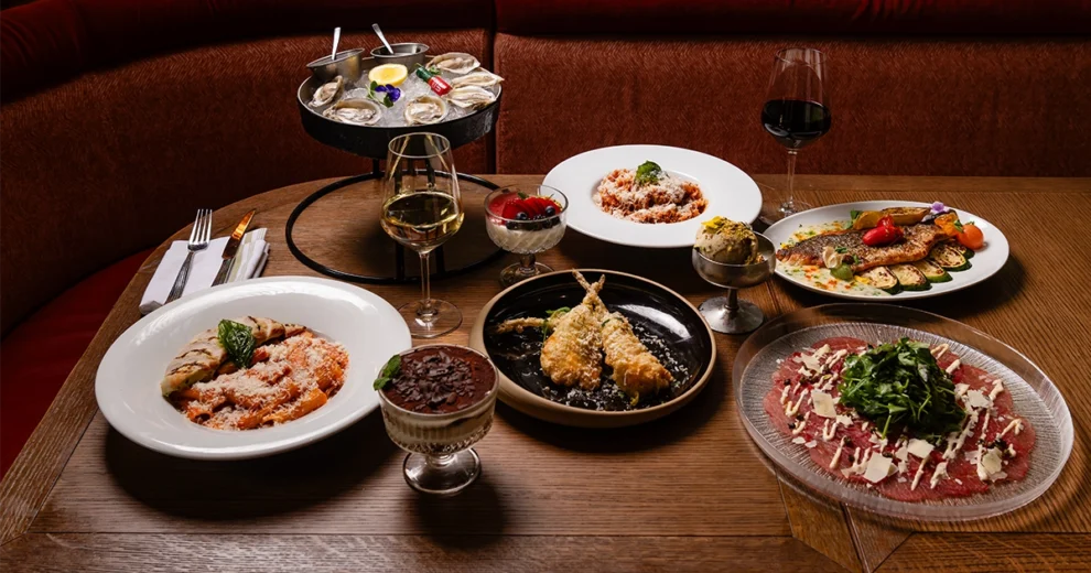 Una mesa de restaurante con una variedad de platos italianos, que incluye pasta, mariscos, carpaccio, fritura de mariscos, verduras, postres, ostras y copas de vino. Los platos están dispuestos sobre una mesa de madera en un comedor de iluminación cálida.