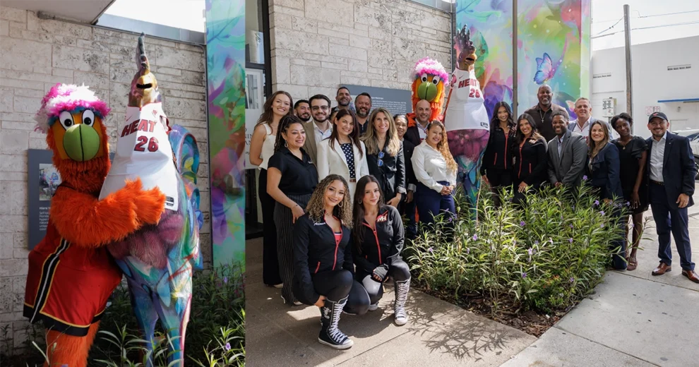 Burnie, la mascota de los Miami HEAT, posa junto a una colorida escultura de un gallo pintado que viste una camiseta del equipo, acompañado por un grupo de empleados de Chase, miembros de la comunidad y bailarines de los HEAT, frente a un edificio decorado con obras de arte vibrantes.