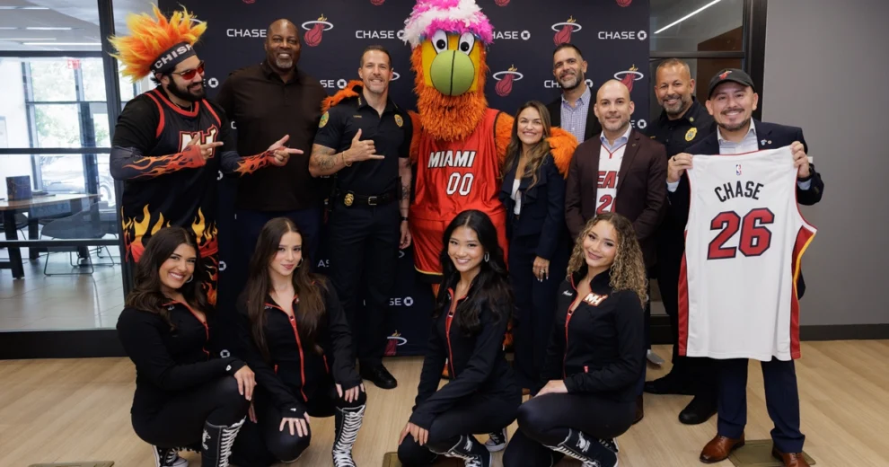 Burnie, la mascota de los Miami HEAT, posa con un grupo de empleados de Chase, agentes del orden y bailarinas de los HEAT dentro de una sucursal de Chase. Una persona sostiene una camiseta blanca de los Miami HEAT con la inscripción "CHASE 26" en la espalda.