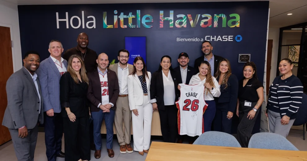 Un grupo de empleados de Chase y representantes de la comunidad posa dentro de una sucursal de Chase, bajo un letrero que dice "Hola Little Havana". Dos personas muestran una camiseta blanca del Miami HEAT firmada y con el número 26.