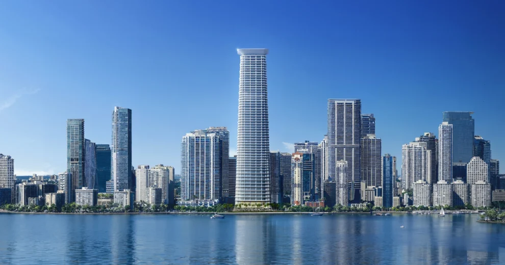 A panoramic daytime view of a modern Miami waterfront skyline, with numerous high-rise residential and office buildings along the water under a clear blue sky. Several boats are visible in the foreground.