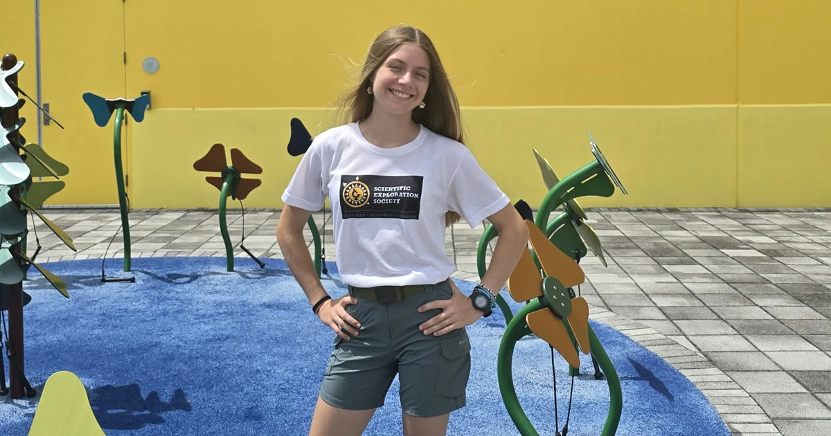 Elvia Gomez smiles outdoors in front of a colorful science-themed play area, wearing a Scientific Exploration Society T-shirt and green shorts.