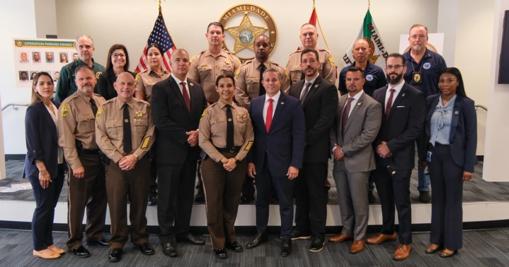 Group photo of Miami-Dade law enforcement officials and community representatives standing in front of the Miami-Dade Sheriff’s Office emblem and U.S., Florida, and Miami-Dade flags. A display board at left features the “Operation Parking Denied” investigation.