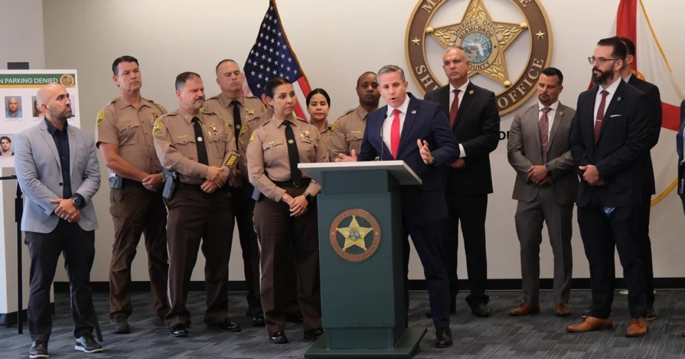El recaudador de impuestos de Miami-Dade, Dariel Fernandez, vestido con traje azul marino y corbata roja, habla desde un podio junto a representantes y funcionarios de las fuerzas del orden de Miami-Dade. Al fondo se aprecian el emblema de la Oficina del Sheriff de Miami-Dade y las banderas de Estados Unidos y Florida, con un panel informativo de la "Operación Estacionamiento Denegado" a la izquierda.