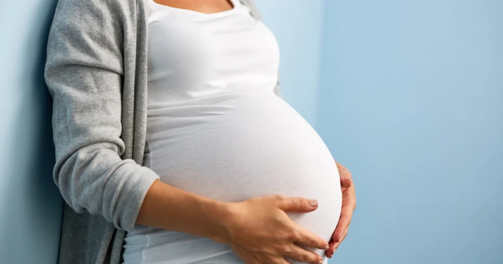 A pregnant woman wearing a white shirt and gray cardigan stands against a light blue background, gently holding her pregnant belly with both hands.