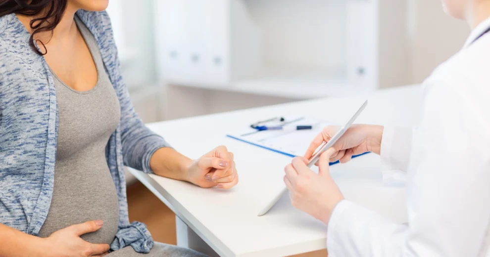 A pregnant woman sits at a desk during a maternal health consultation with a healthcare professional, who is reviewing information on a tablet. The woman rests one hand on her abdomen.