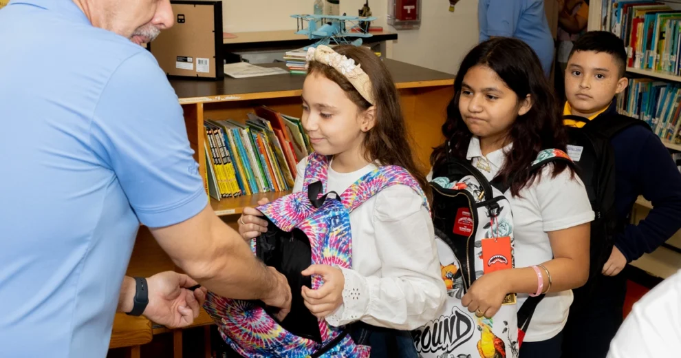 Elementary school students stand in line to receive backpacks during a back-to-school event hosted by the Kiwanis of Little Havana Foundation in partnership with the Miami Marlins Foundation. A volunteer hands a colorful backpack to one student while two others wait their turn with backpacks on their shoulders.
