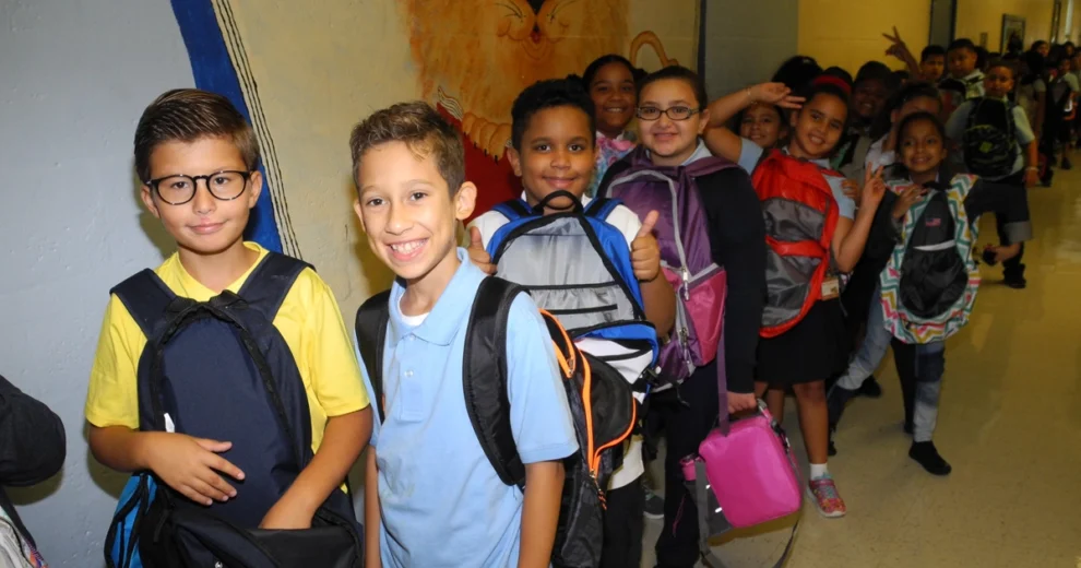 Alumnos de primaria con mochilas coloridas hacen fila en un pasillo de la escuela durante un evento de regreso a clases organizado por la Fundación Kiwanis de Little Havana en colaboración con la Fundación Miami Marlins. Varios niños sonríen a la cámara mientras se preparan para el nuevo año escolar.