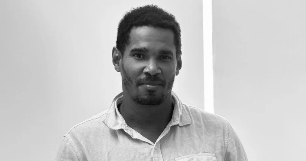 Black-and-white portrait of Cuban artist and activist Luis Manuel Otero Alcántara looking directly at the camera indoors. He has short curly hair, a trimmed beard, and wears a light-colored button-up shirt. The background features a simple white wall, keeping the focus on his face and expression.