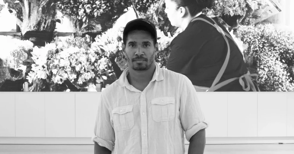 Black-and-white photograph of Cuban artist and activist Luis Manuel Otero Alcántara standing in front of a large floral-themed artwork.