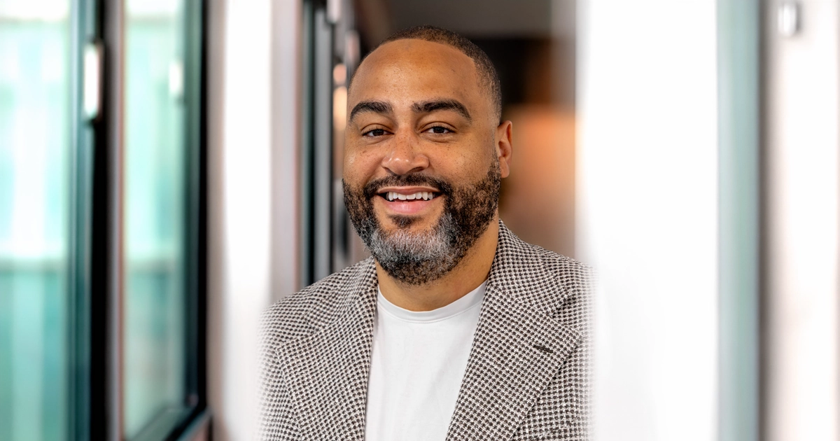 MJ Green of the Miami Downtown Development Authority smiles at the camera while standing in a bright, modern indoor setting. He is wearing a patterned blazer over a white shirt.