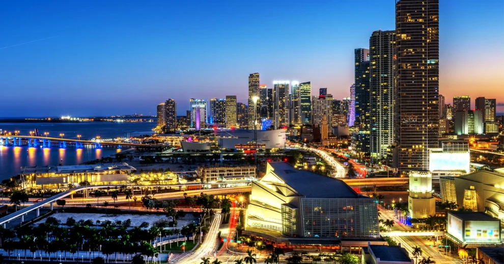 A panoramic nighttime view of downtown Miami, Florida, featuring illuminated skyscrapers, busy roads, bridges, waterfront areas and buildings beneath a blue and orange evening sky.