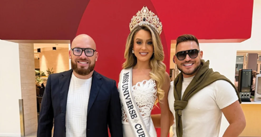 Miss Universe Cuba Mia Dio poses wearing her crown and sash between two men inside a hotel or event venue.