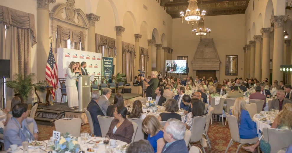 Attendees gather for the SFLHCC Sustainability Conference in an ornate ballroom as a speaker addresses the audience from a podium. The stage features an American flag, South Florida Hispanic Chamber of Commerce banners and event signage.