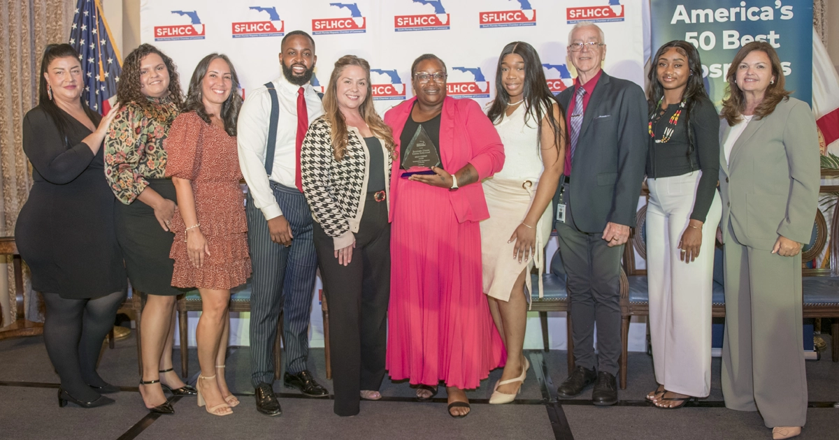 A group of nine people poses during the SFLHCC Sustainability Conference, with a woman in a bright pink dress holding an award. South Florida Hispanic Chamber of Commerce banners and an American flag are visible in the background.