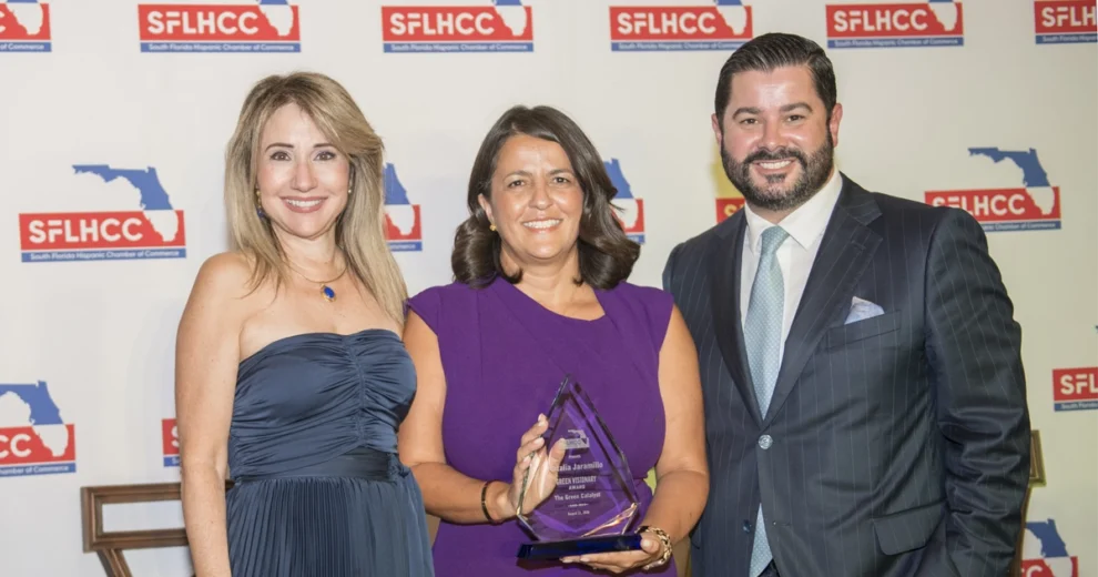 Three people pose at the SFLHCC Sustainability Conference in front of South Florida Hispanic Chamber of Commerce banners. The woman in the center holds a glass award recognizing her with the Green Visionary Award.