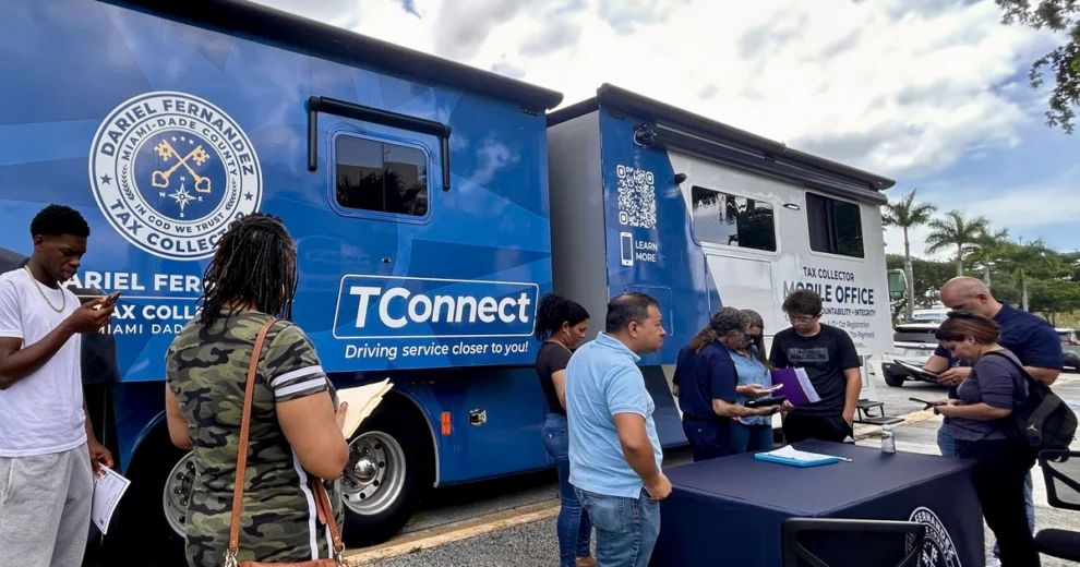 La gente se reúne alrededor de la oficina móvil TConnect de la oficina de recaudación de impuestos de Miami-Dade, estacionada al aire libre. Varios residentes están de pie junto a una mesa de registro, mientras otros completan trámites o utilizan sus teléfonos.