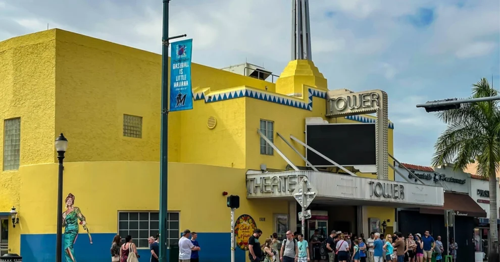 Vista exterior diurna del histórico "Tower Theater", en la Calle Ocho de Little Havana, Miami. El edificio, de color amarillo brillante y estilo Art Déco, presenta detalles ornamentales geométricos en azul y blanco, el destacado letrero luminoso con el nombre TOWER THEATER y una multitud de personas reunidas en el exterior.