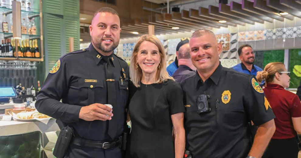 Miami-Dade County Commissioner Vicki Lopez stands smiling between two City of Miami Police officers inside a restaurant during a community event. The officers are in full uniform, and other attendees can be seen socializing in the background.