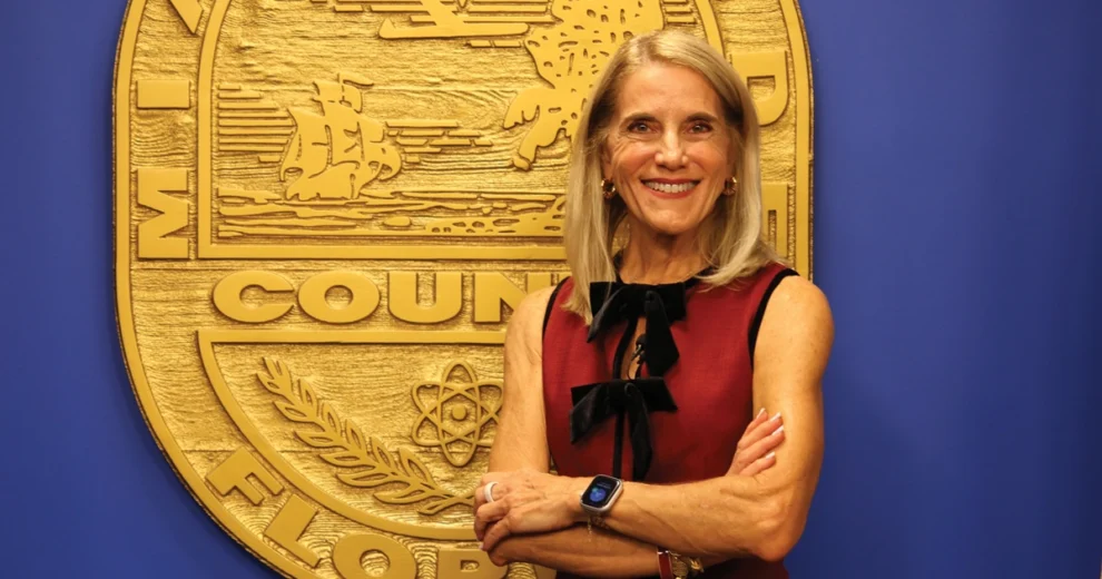 La comisionada del condado de Miami-Dade, Vicki Lopez, posa con los brazos cruzados frente al sello del condado de Miami-Dade, colocado sobre una pared azul.