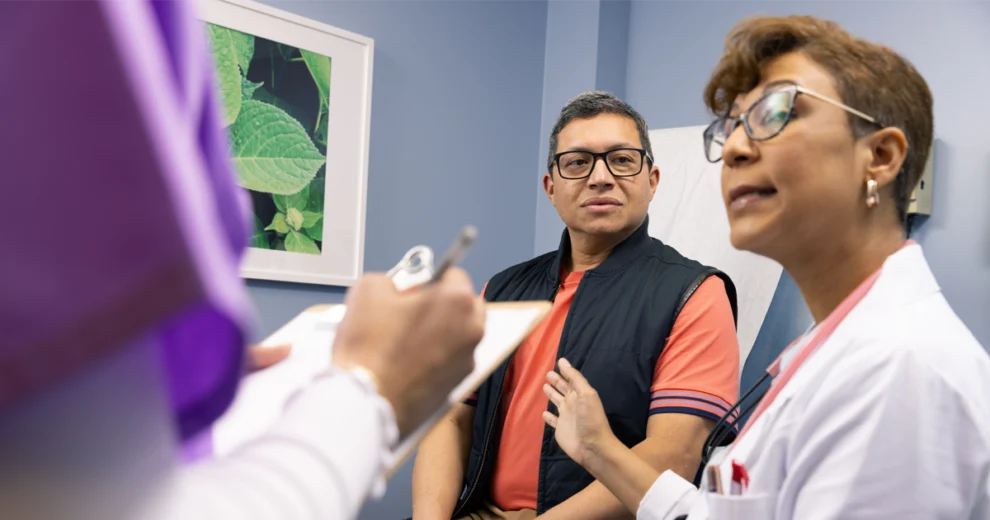 A healthcare professional discusses cholesterol health with a male patient during a medical appointment, while another healthcare worker takes notes, highlighting the importance of monitoring and managing cholesterol.