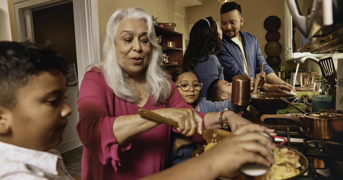 Una familia de varias generaciones prepara alimentos junta en la cocina, destacando la alimentaci&oacute;n saludable y la importancia de controlar el colesterol como parte de la salud cardiovascular general.