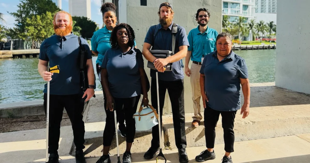 A group of six people poses outdoors beside a waterfront, with several wearing matching polo shirts and holding trash grabbers. Buildings and water are visible in the background.