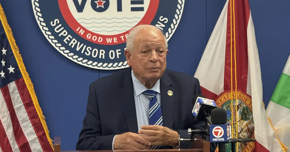A man in a blue suit speaks at a podium during a press conference, with U.S., Florida, and Miami-Dade County flags and the Supervisor of Elections seal behind her.