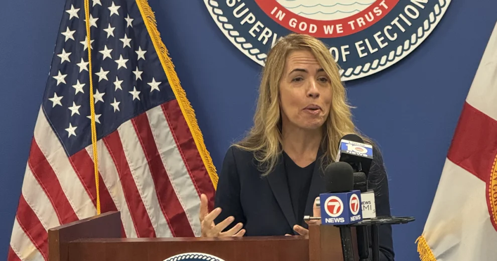 Miami-Dade Commissioner Raquel Regalado speaks about accessible voting at a podium during a press conference at the Miami-Dade County Supervisor of Elections office, with U.S. and Florida flags and the Supervisor of Elections seal visible behind her. Multiple news microphones are positioned in front of her.