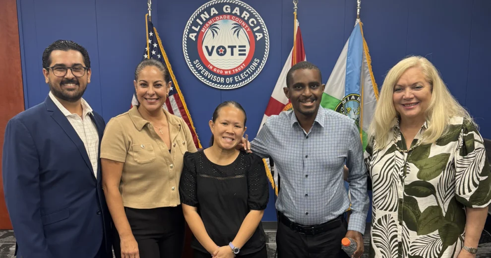 Five people pose for a group photo inside the Miami-Dade County Supervisor of Elections office, standing in front of the official seal and American, Florida, and Miami-Dade County flags.