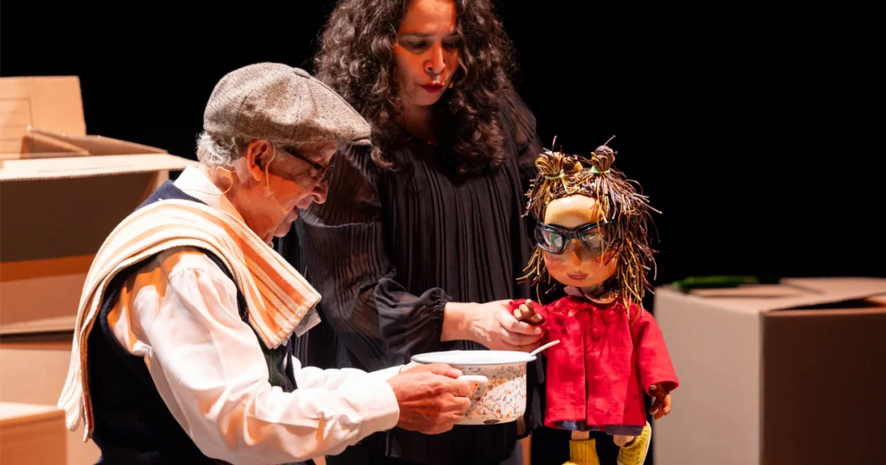 A theatrical performance from the Adrienne Arsht Center’s Hispanic programming featuring an older man holding a bowl beside a woman and a puppet dressed in a red coat and glasses.